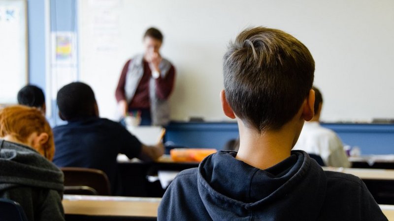 Ein Schüler von hinten dargestellt, der in einem Klassenzimmer sitzt und richtung Lehrperson und Tafel schaut