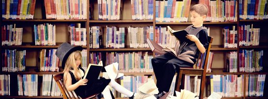 Kinder sitzen auf hohen Stühlen begeistert in einer Bibliothek und schmökern und stöbern in Büchern.