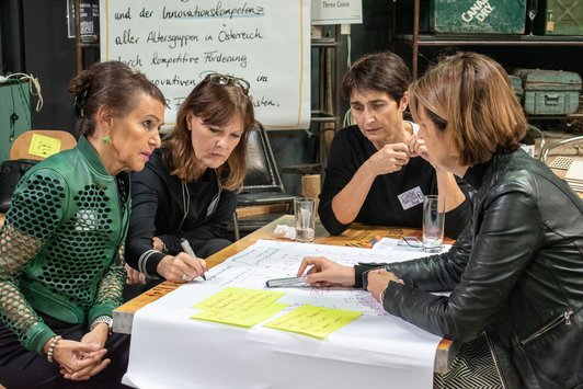 Teilnehmerinnen sitzen als Gruppe auf einem alten Schultisch und arbeiten zusammen