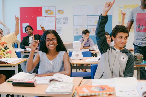 Schüler*innen in einem Klassenzimmer, die aufzeigen. In ihrer Mitte der Avatar AV1.